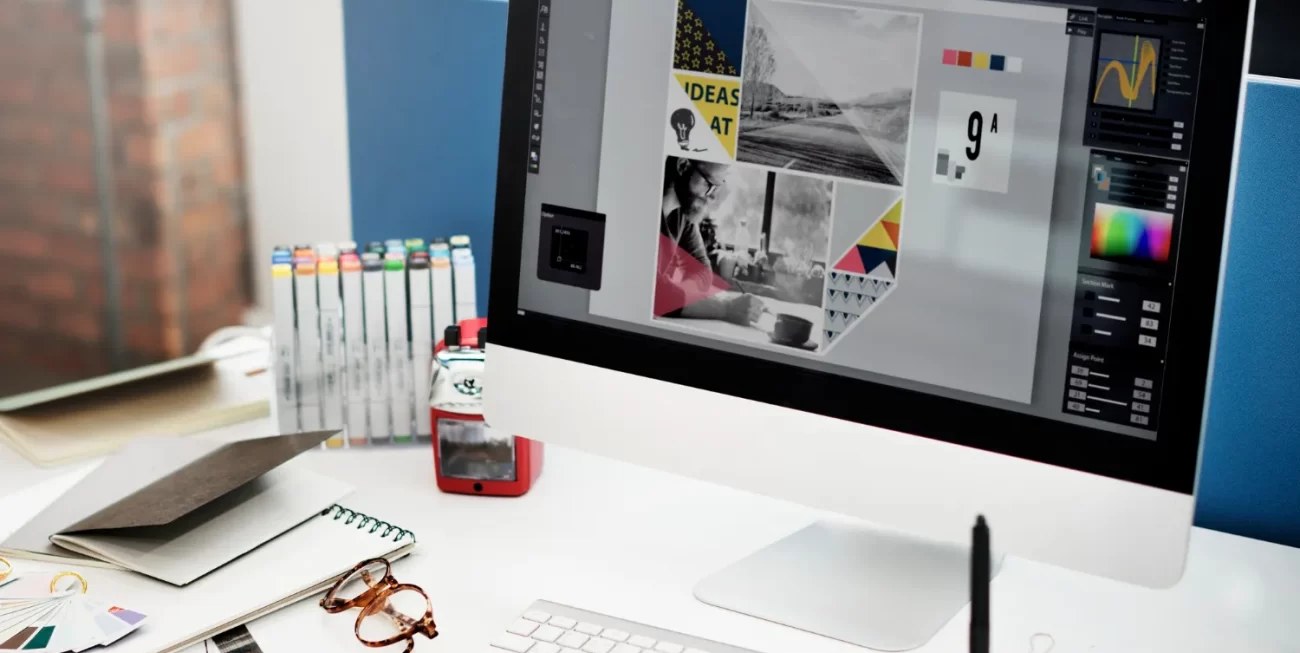 Ordenador de escritorio con pantalla mostrando un diseño gráfico, rodeado de herramientas de trabajo como marcadores, cuadernos y gafas, en un entorno de oficina creativa.