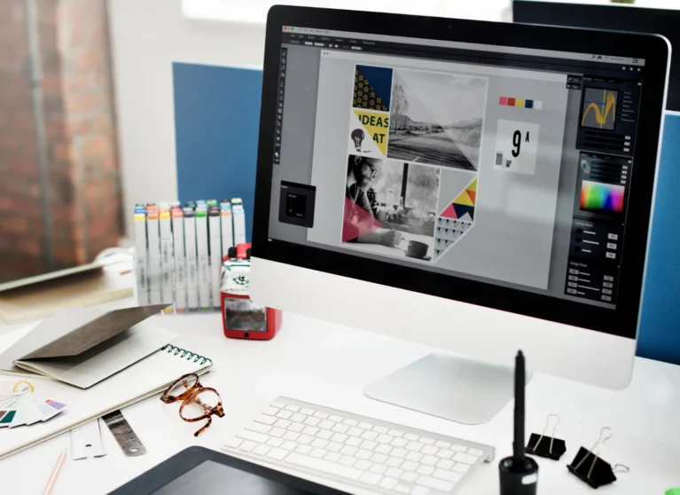 Ordenador de escritorio con pantalla mostrando un diseño gráfico, rodeado de herramientas de trabajo como marcadores, cuadernos y gafas, en un entorno de oficina creativa.