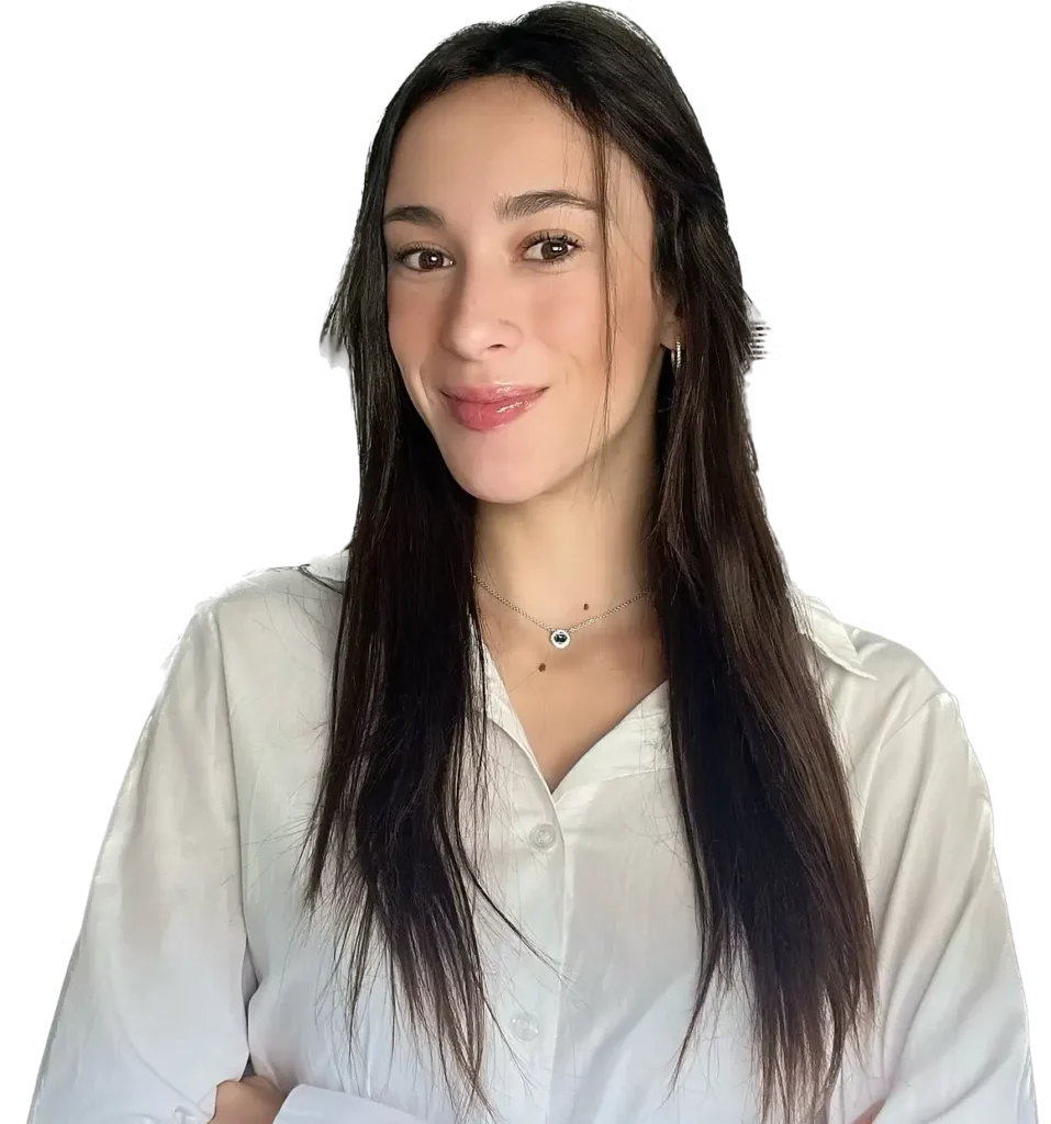 Mujer joven con cabello largo y oscuro, vestida con una camisa blanca, sonriendo y posando con los brazos cruzados. Fondo negro.